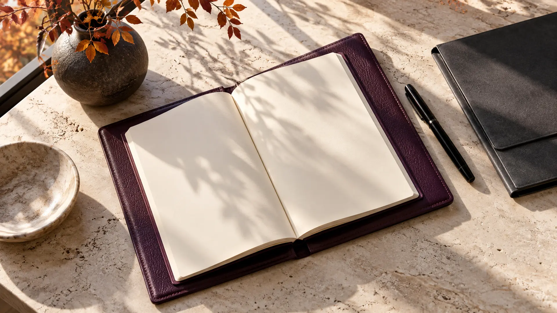 An open plum leather planner with blank pages, a pen, and autumn foliage on a stone desk.